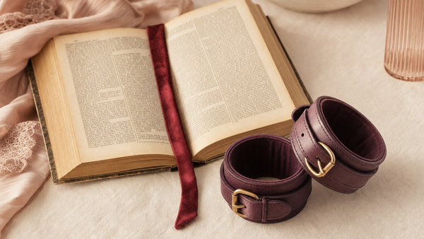 Dizionario aperto su un tavolo avorio con segnalibro in raso bordeaux e un paio di polsiere in pelle color prugna
