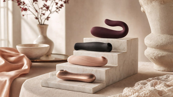 Quattro oggetti di design in silicone prugna, nero, cipria e oro rosa su un espositore di marmo a gradini